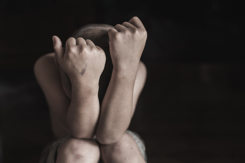 A child with a shaved head sits alone, arms crossed over their face defensively. The dimly lit setting conveys a mood of vulnerability and sadness.